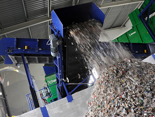 Blue industrial conveyor system dumping shredded recyclable material into a large pile inside a processing facility.