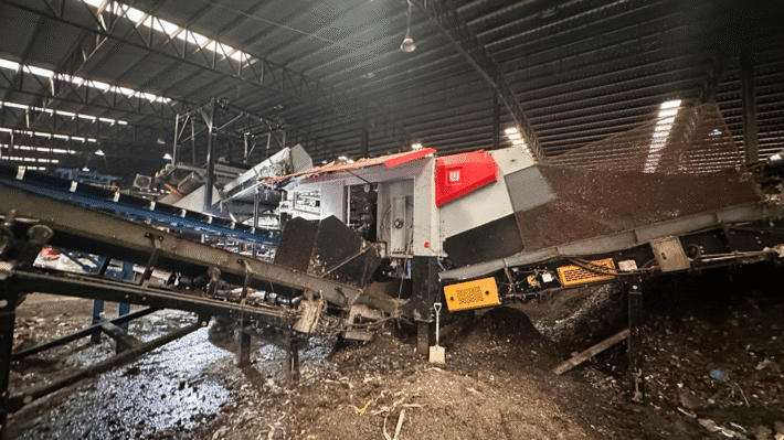 Industrial sorting machine inside a dim processing facility, with conveyor belts moving debris and material scattered on the floor.