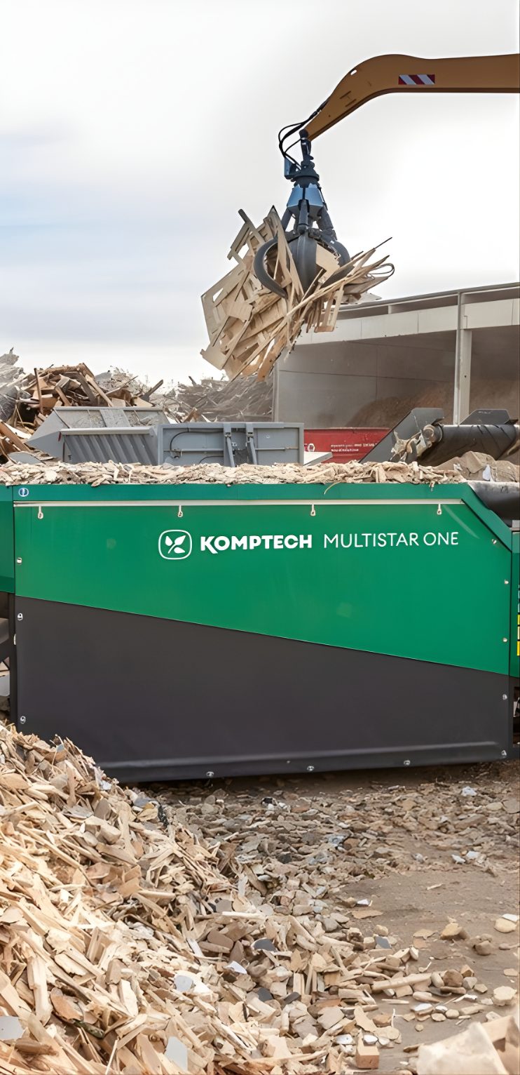 Wood waste grinder processing scrap wood beside an excavator at a recycling yard.