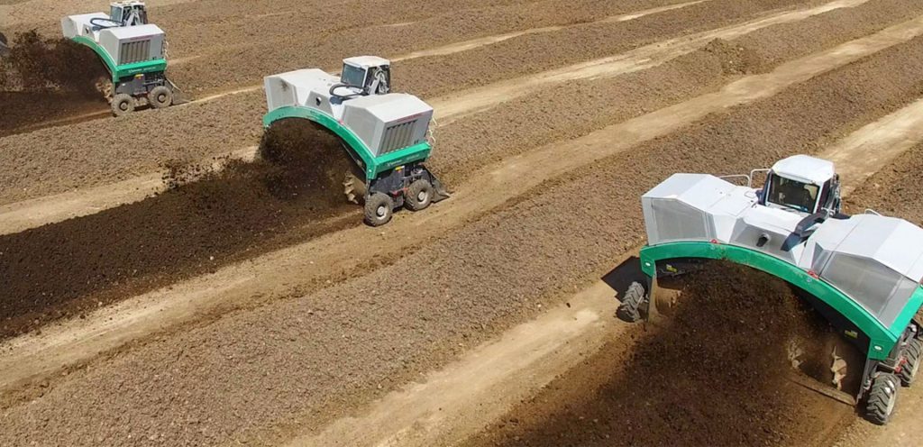 Three large green and white compost turner machines working across long rows of soil or compost in an open field.